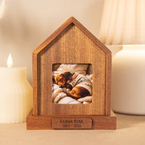 Może przedstawiać: Drewniany pomnik dla zwierząt w kształcie domu ze zdjęciem śpiącego psa w swetrze. Pomnik ma prostokątną podstawę z napisem "LUNA KIM 2012-2023". Świeca i lampa są w tle.