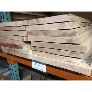 Puede incluir: Una pila de tablas de madera de 10 pulgadas de grosor, secadas al aire. Las tablas son de color marrón claro y tienen una superficie lisa.