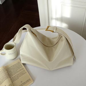 Könnte beinhalten: Cremefarbene Umhängetasche mit goldenem Reißverschluss und Schnalle. Die Tasche hat einen breiten Riemen und eine geschwungene Form. Ein weißer Becher und ein offenes Buch liegen ebenfalls auf dem Tisch.