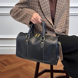 以下が含まれることがあります： ゴールドカラーの金具（ジッパーやリベットなど）が付いた黒い革製のダッフルバッグ。バッグには2つのトップハンドルとショルダーストラップアタッチメントが付いています。バッグは長方形で、底がカーブしており、白いステッチが施されています。