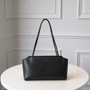 Könnte beinhalten: Eine einfarbige schwarze Schultertasche mit rechteckigem Boden und einer glatten, matten Oberfläche. Die Tasche verfügt über zwei dünne, vertikale Trageriemen an der Oberkante, die in der Mitte der Öffnung ein leicht geschwungenes Design aufweist.
