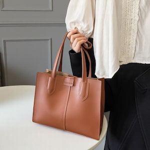 Könnte beinhalten: Eine braune Ledertasche mit zwei Henkeln. Die Tasche hat eine rechteckige Form mit einer vertikalen Naht in der Mitte und dem Wort "FASHION" auf einem kleinen Etikett. Die Tasche wird von einer Person gehalten.