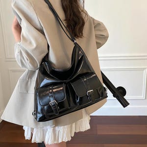 May include: A black shoulder bag with two front pockets, each secured with a buckle closure. The bag has a glossy finish and a long strap. The bag is worn over the shoulder.