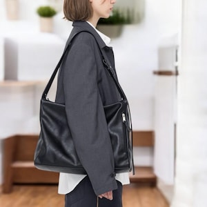 May include: A black leather messenger bag with a long shoulder strap. The bag has a rectangular shape with a zipper pocket on the front. The bag is worn over a gray blazer and white shirt.