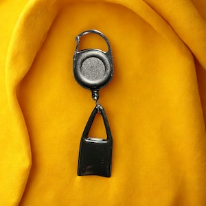 Puede incluir: Un carrete retráctil de insignia negro con un clip plateado y una etiqueta rectangular negra. El carrete está unido a una etiqueta negra, todo sobre un fondo amarillo.