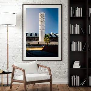 Può includere: Fotografia incorniciata di un'alta struttura architettonica bianca con un intricato design traforato su uno sfondo di cielo blu chiaro. L'immagine è ambientata in una stanza con un muro di mattoni bianchi, pavimento in legno e una sedia moderna.