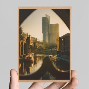 May include: A postcard featuring a photograph of a canal in a city with tall buildings in the background. The canal is lined with brick buildings and a bridge. There are boats in the water. The photo is framed with a brown border.