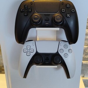Könnte beinhalten: Eine weiße und schwarze Spielkonsole mit zwei Controllern. Der obere Controller ist schwarz und der untere Controller ist weiß. Die Controller sind auf einem weißen Ständer montiert. Die Konsole ist weiß mit einem schwarzen Sockel.
