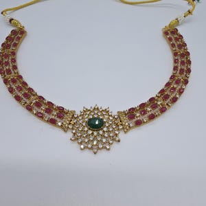 May include: A gold-toned necklace featuring three rows of oval-shaped red stones on each side. The center piece is a circular sunburst design set with small clear stones surrounding a single oval green stone in the middle.