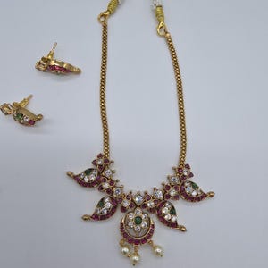 May include: A gold-toned necklace and matching earring set featuring teardrop-shaped motifs inlaid with red and green stones and clear crystals. The necklace includes a central floral pendant with three dangling white beads.
