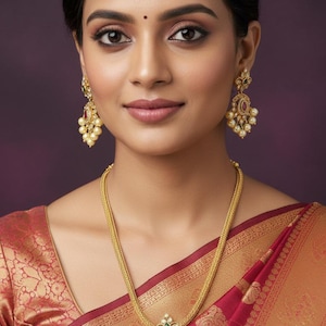 May include: A woman wearing gold jewelry, including earrings and a necklace with a pendant. The pendant features a central red stone surrounded by smaller white stones and pearls. The woman is wearing a red and gold sari.