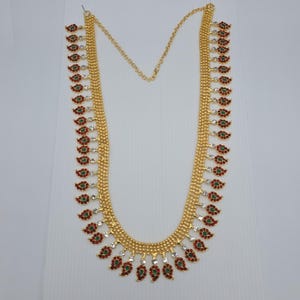 May include: A long gold-tone necklace featuring a series of dangling paisley-shaped charms. Each charm is accented with small red and green stones, interspersed with clear white crystals along the length of the chain, creating a decorative pattern.