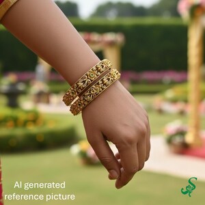 May include: Gold-colored bangles with intricate floral and scrollwork designs, adorned with small green and clear gemstones. The bangles are worn on a wrist, showcasing their ornate details. The background is a blurred outdoor scene.