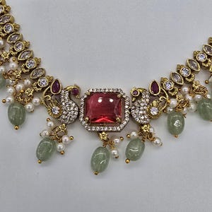 May include: A gold-toned necklace featuring a central rectangular red stone surrounded by clear crystals. Two peacock motifs with clear crystals and red accents flank the center. The piece is adorned with small white pearls and pale green oval beads.