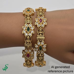 May include: Two gold-colored bangles with intricate floral and figure designs. Each bangle features a series of flower-shaped settings with clear and colored gemstones. The bangles are adorned with small statues and are displayed on a light background.