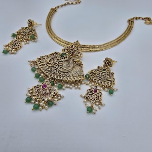 May include: A gold-toned necklace and matching earring set featuring intricate filigree metalwork, embedded with small clear stones, teardrop-shaped pink stones, and round green beads, accented with clusters of tiny white pearls hanging from the base.