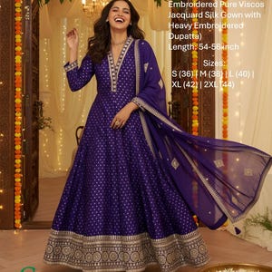 May include: A long, flowing, dark purple gown with gold embroidered details and a matching dupatta. The dress features a V-neckline and a full skirt. The text on the image says "Fabric Detail Pure Viscos Jacquard Silk Gown with Heavy Embroidered Dupatta".