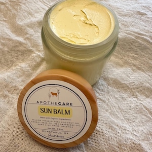May include: A jar of yellow sun balm with the lid off, next to the wooden lid. The label reads "SUN BALM" and lists ingredients. The jar contains 3.3 oz of product.