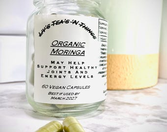 May include: A glass jar with a white label that reads "Liv's Tea's 'N Things Organic Moringa May Help Support Healthy Joints and Energy Levels 60 Vegan Capsules Best If Used By March 2027". Two green capsules are in front of the jar.