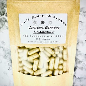 May include: A kraft paper bag with a clear window, filled with oblong capsules. The label reads "Liv's Tea's 'n Things" and "Organic German Chamomile." The bag contains 120 capsules, each with 350+ mg. The text "Best if used by June 2028" is also visible.