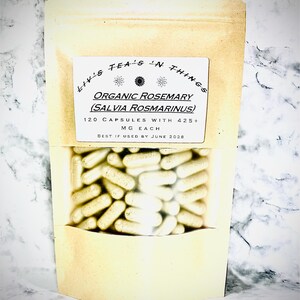 Op de afbeelding: Een bruin papieren zakje met een transparant venster dat talrijke witte capsules toont. Het etiket vermeldt "Liv's Tea's 'N Things" en "Organic Rosemary (Salvia Rosmarinus)". Het geeft ook aan "120 Capsules met 425+ MG per stuk" en "Ten minste houdbaar tot juni 2028."