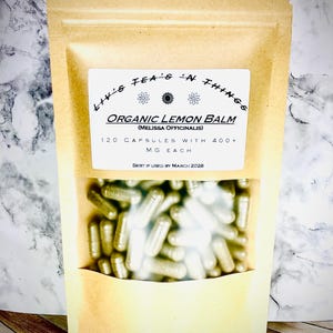 May include: A brown paper bag with a label that reads "ttt's TEA'S 'N THINGS ORGANIC LEMON BALM (MELISSA OFFICINALIS) 120 CAPSULES WITH 400+ MG EACH BEST IF USED BY MARCH 2028". The bag contains a clear plastic bag filled with silver capsules.