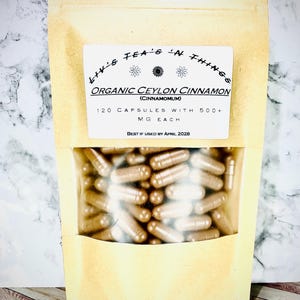 May include: A tan pouch with a clear window displaying numerous brown capsules. The pouch has a white label with the text "Liv's Tea's 'n Things" and "Organic Ceylon Cinnamon (Cinnamomum)". It also states "120 Capsules with 500+ MG Each" and "Best if Used by April 2028".