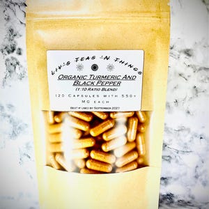Op de afbeelding: Een bruin zakje met een doorzichtig venster met talrijke oranje capsules. Het etiket luidt "Liv's Teas 'n Things" en "Organic Turmeric and Black Pepper". De tekst geeft ook 120 capsules met 550+ mg aan, met een houdbaarheidsdatum van september 2027.