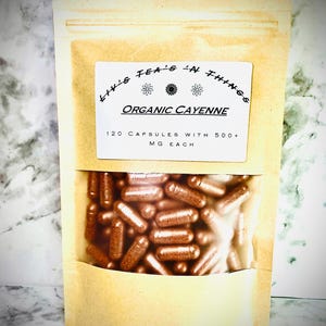 May include: A tan paper bag with a clear window displaying numerous brown capsules. The bag has a white label with the text "Liv's Tea's 'n Things" and "Organic Cayenne". The label also states "120 Capsules with 500+ MG Each".