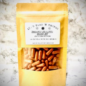 Peut inclure: Un sachet debout beige contenant 100 gélules de cayenne bio 60 000 HU. L'étiquette indique : Liv's Teas 'N Things, Organic Cayenne 60 000 HU (Capsicum Annuum), 100 gélules de 500+ mg chacune, à consommer de préférence avant septembre 2026.
