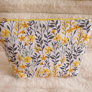 May include: A white cosmetic bag with a floral pattern of yellow flowers and gray leaves. The bag has a yellow zipper and a rectangular shape, perfect for storing makeup or toiletries.