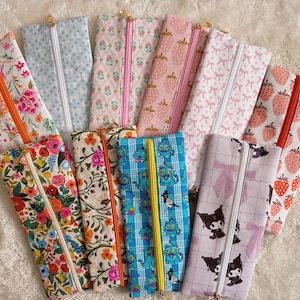 May include: A collection of pencil cases with various patterns, including floral, strawberry, and bow designs. The cases have different colored zippers, such as orange, pink, and yellow. The cases are approximately 8 inches long.