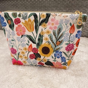 May include: A floral print cosmetic bag featuring a vibrant array of flowers including sunflowers, tulips, and roses. The bag has a gold zipper and a white background. The design includes ladybugs and various green leaves.