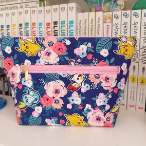 May include: A navy blue zippered pouch with a floral and cartoon character pattern. The pouch features a pink zipper and gold-colored zipper pull. The design includes pink and white flowers and yellow cartoon characters.