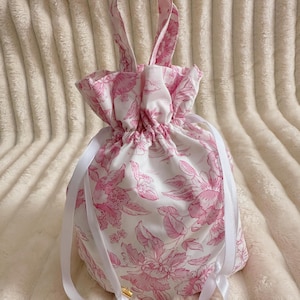 Puede incluir: Una bolsa blanca con cordón y estampado floral rosa. La bolsa tiene una parte superior fruncida, lazos de cinta blanca y asas a juego. Los detalles metálicos dorados están en la base de los lazos. La bolsa está sobre una superficie texturizada color crema.