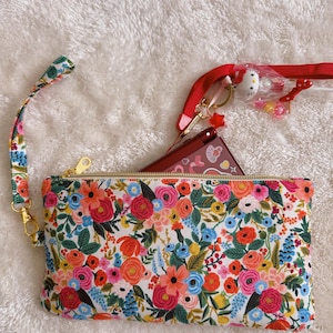 May include: A floral print clutch with a gold zipper, a wrist strap, and a red lanyard with a Hello Kitty charm. The clutch features a colorful pattern of flowers in shades of pink, orange, blue, and green on a white background.
