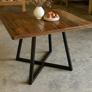 May include: A square wooden table with a dark brown finish, supported by black metal legs. A white vase with dried flowers and a small bowl of fruit sit on the tabletop. The table's dimensions are approximately 91 cm square.