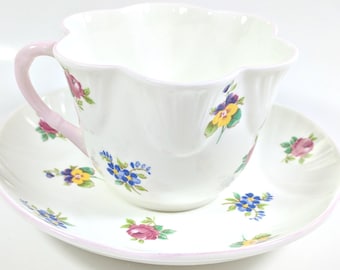 May include: A white teacup and saucer set with a pink rim and floral pattern. The cup has a scalloped edge and a pink handle. The saucer has a scalloped edge and a floral pattern.