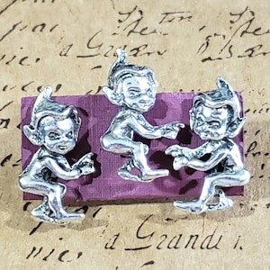 以下が含まれることがあります： 紫色の表面に、それぞれ異なるポーズをとった銀製のエルフの置物が3つあります。エルフはとがった耳と大きな目を持ち、シンプルな服を着ています。