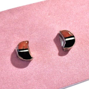 Könnte beinhalten: Ein Paar silberfarbene Ohrstecker mit einem einzigartigen Design. Jeder Ohrring hat eine gebogene Form mit Abschnitten in Orange, Schwarz und Weiß, vor einem rosa Hintergrund. Die Ohrringe haben eine moderne, geometrische Ästhetik.