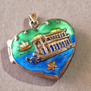 May include: A heart-shaped locket pendant with a gold-colored metal frame and enamel detailing. The locket features a scene of a building on a body of water with green and blue enamel. The locket has a hinged opening.