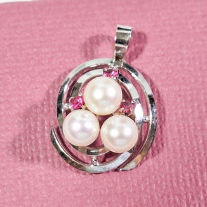 May include: A silver pendant featuring three large, round pearls and small pink gemstones. The pendant has a spiral design around the pearls and a loop at the top for a chain. The background is a solid pink colour.