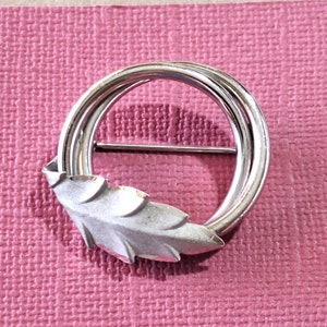 May include: A silver tone brooch with a circular design and a leaf detail. The brooch is on a pink background.