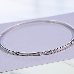 May include: A silver bangle bracelet with a delicate, engraved design. The bracelet features a repeating pattern of curved lines and small shapes. The thin, circular band is displayed on a light purple surface.