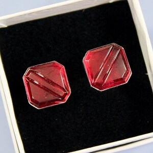 May include: A pair of red, square-shaped earrings with diagonal lines, presented in a white box with a black interior. The earrings have a faceted design and a silver-colored border, perfect for adding a touch of elegance.