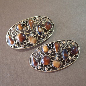 May include: Two oval brooches with a gold-coloured frame. Each brooch has a filigree design and is adorned with brown, amber, and clear gemstones of various shapes. The gemstones are set within the gold frame, creating a decorative accessory.