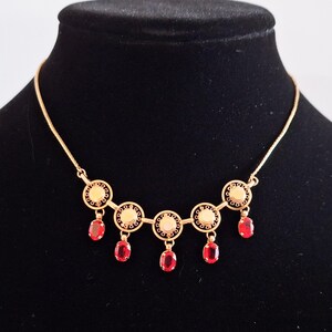 May include: A gold-toned necklace with a series of circular pendants and dangling red gemstones. The necklace is displayed on a black mannequin, showcasing its design and color contrast. The necklace features intricate detailing.