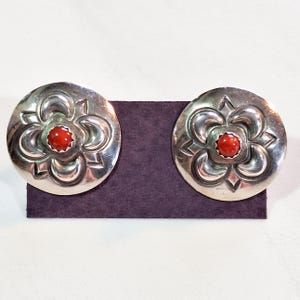 May include: Pair of silver-toned stud earrings. Each earring features a floral design with a central red stone. The earrings are displayed on a dark purple surface against a white background. The earrings have a vintage or Southwestern style.