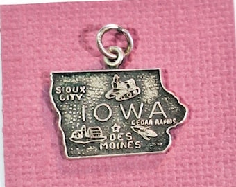 Könnte beinhalten: Silberfarbener Anhänger in Form des Bundesstaates Iowa mit dem Wort "Iowa" und den Städten Sioux City, Cedar Rapids und Des Moines. Der Anhänger hat eine Schlaufe zum Befestigen an einer Halskette oder einem Armband. Der Anhänger befindet sich auf einem rosa Hintergrund.