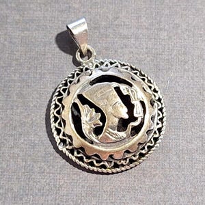 May include: A silver pendant with a detailed design. The pendant features a central image of a profile, possibly of a historical figure, surrounded by an ornate border. The pendant has a loop for a chain.